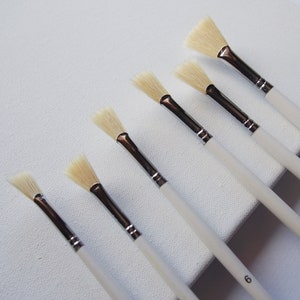 May include: Set of seven white handled fan paint brushes with white bristles. The brushes are arranged in order of size, with the largest brush at the top and the smallest brush at the bottom.