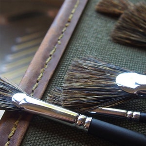 May include: A close-up of a set of paint brushes with natural bristles and silver metal ferrules. The brushes are arranged on a canvas-like surface with a brown leather strap.