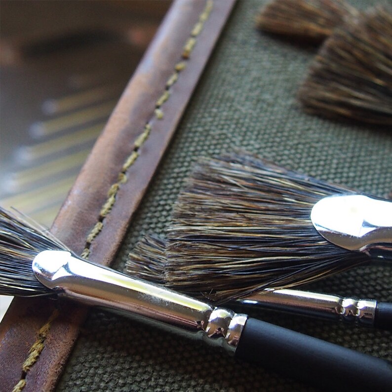 May include: A close-up of a set of paint brushes with natural bristles and silver metal ferrules. The brushes are arranged on a canvas-like surface with a brown leather strap.