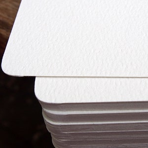 May include: Close-up of a stack of white, textured cards with rounded corners. The top card is in focus, revealing the paper's subtle grain. The cards are stacked neatly, suggesting a stationery or paper product.
