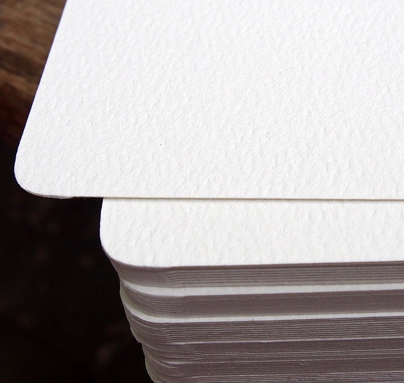 May include: Close-up of a stack of white, textured cards with rounded corners. The top card is in focus, revealing the paper's subtle grain. The cards are stacked neatly, suggesting a stationery or paper product.