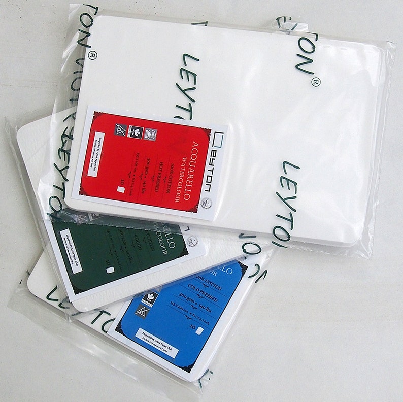 May include: Several packages of Leyton watercolor paper are displayed. Each package contains white paper and a label with details like "ACQUARELLO WATERCOLOUR" and "100% COTTON." The labels are red, green, and blue, indicating different paper types.