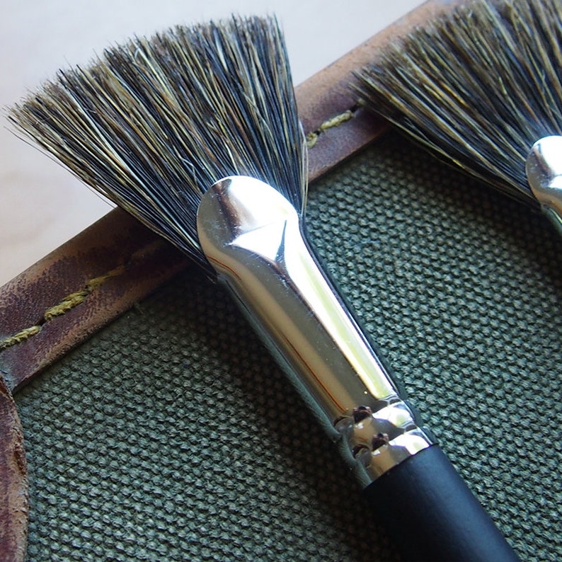 May include: A close-up of a fan brush with black and silver bristles. The brush is in a leather case with a green canvas lining.