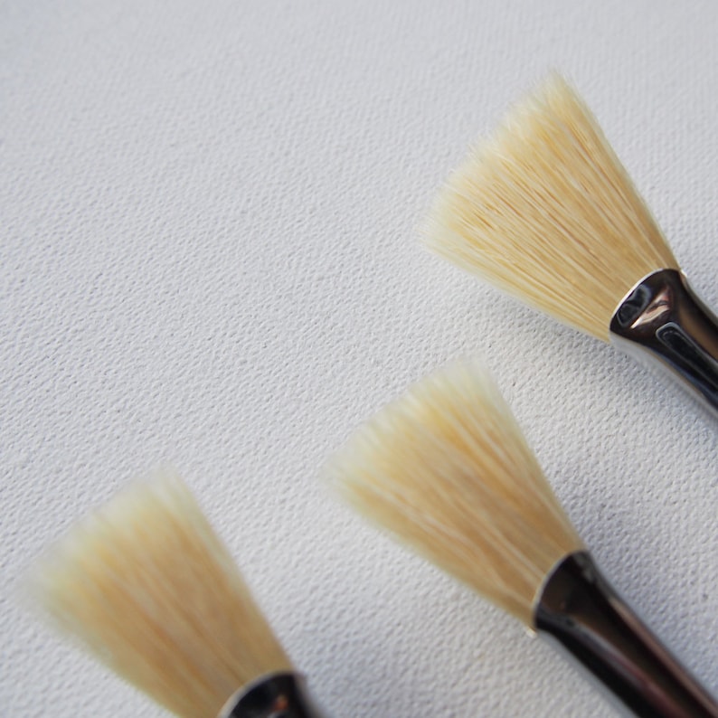 May include: Three paint brushes with natural bristles and silver handles. The brushes are arranged in a row on a white surface.