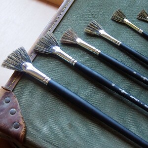 May include: Set of five fan paint brushes with black handles and silver ferrules. The brushes are arranged in order of size, with the largest brush at the bottom and the smallest brush at the top. The brushes are labeled with their size in white lettering on the handles. The largest brush is labeled "10", the next largest is labeled "8", and the remaining brushes are labeled "6", "4", and "2".