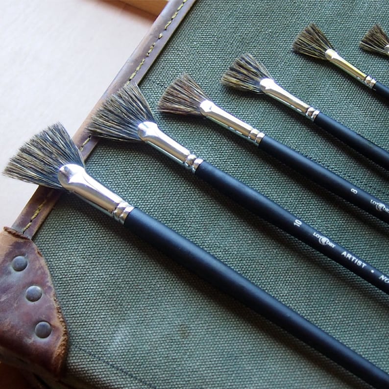May include: Set of five fan paint brushes with black handles and silver ferrules. The brushes are arranged in order of size, with the largest brush at the bottom and the smallest brush at the top. The brushes are labeled with their size in white lettering on the handles. The largest brush is labeled "10", the next largest is labeled "8", and the remaining brushes are labeled "6", "4", and "2".