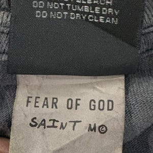 May include: A black clothing care label featuring white text that reads DO NOT IRON DECORATION, USE PRESS CLOTH, DO NOT BLEACH, DO NOT TUMBLE DRY, DO NOT DRY CLEAN. Below is a white tag with FEAR OF GOD SAINT M, SIZE OS, and BODY 100% COTTON.