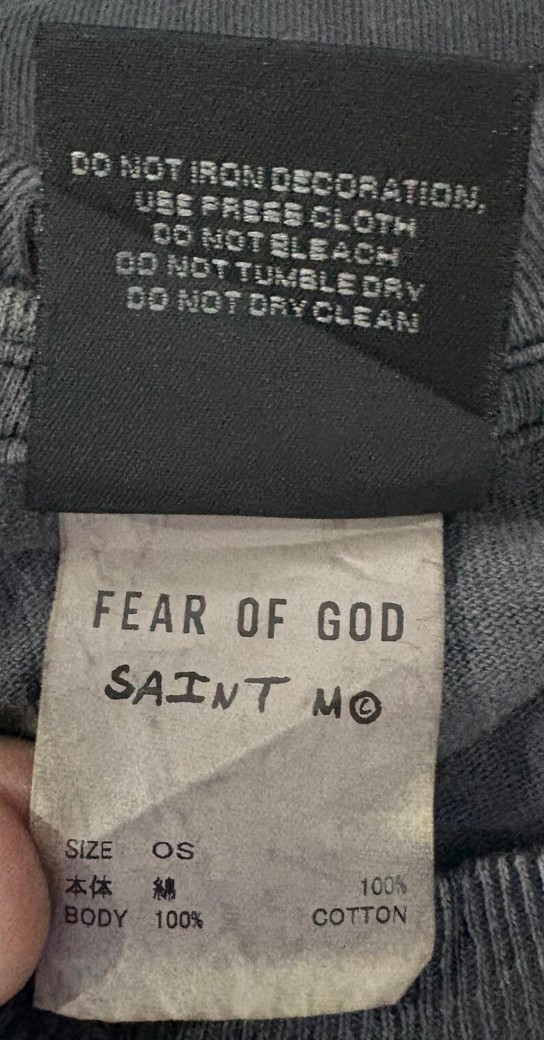 May include: A black clothing care label featuring white text that reads DO NOT IRON DECORATION, USE PRESS CLOTH, DO NOT BLEACH, DO NOT TUMBLE DRY, DO NOT DRY CLEAN. Below is a white tag with FEAR OF GOD SAINT M, SIZE OS, and BODY 100% COTTON.