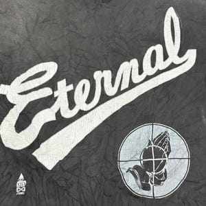 May include: A black fabric garment featuring the white text Eternal in a large script font. Below the text is a circular graphic containing a crosshair and a pair of praying hands. A small triangular logo with the text Saint of Eden is visible.