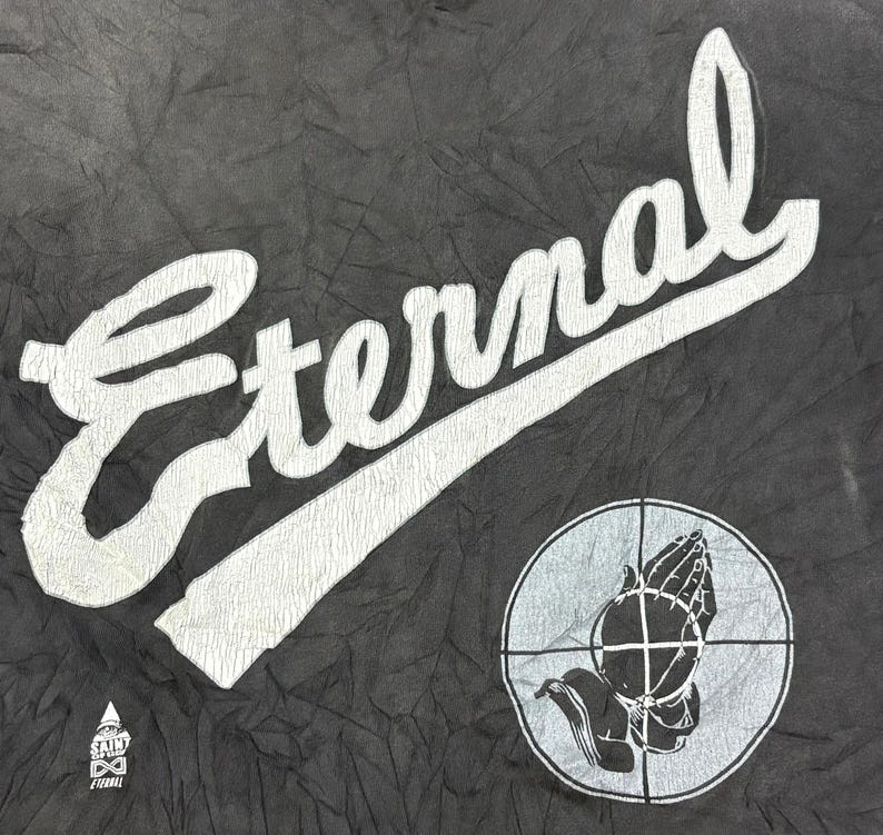 May include: A black fabric garment featuring the white text Eternal in a large script font. Below the text is a circular graphic containing a crosshair and a pair of praying hands. A small triangular logo with the text Saint of Eden is visible.