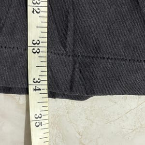 May include: A dark gray fabric garment edge featuring a double-stitched hemline. A white measuring tape is positioned vertically against the fabric, with the tape showing numerical markings from 32 to 35 inches along the edge of the material.