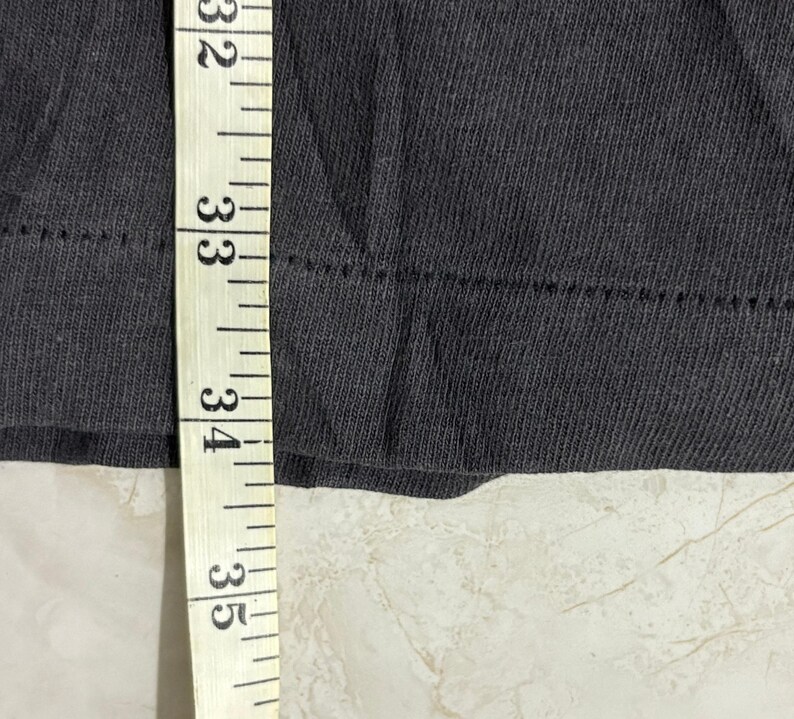 May include: A dark gray fabric garment edge featuring a double-stitched hemline. A white measuring tape is positioned vertically against the fabric, with the tape showing numerical markings from 32 to 35 inches along the edge of the material.