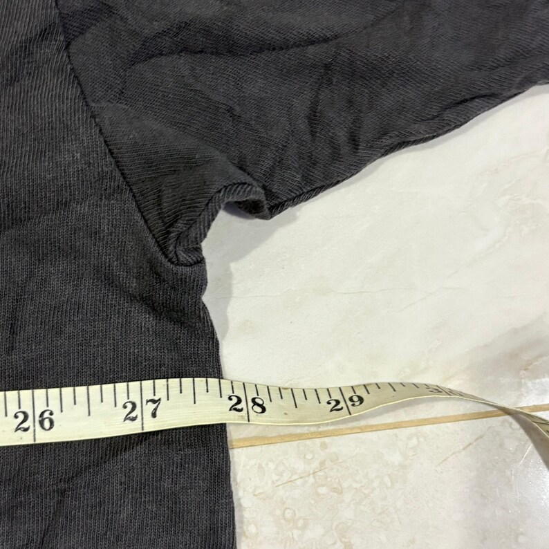 May include: A dark gray t-shirt featuring a visible seam at the armhole and side. A yellow measuring tape is positioned horizontally across the fabric, showing markings from 26 to 29 inches to indicate the width of the garment.