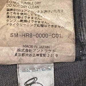 May include: A rectangular fabric care label with black text on a light background. It includes laundry symbols, instructions like DO NOT IRON DECORATION, the code SM-HR8-0000-C01, and MADE IN JAPAN. Below is a small white tag with a black line drawing of an angel.
