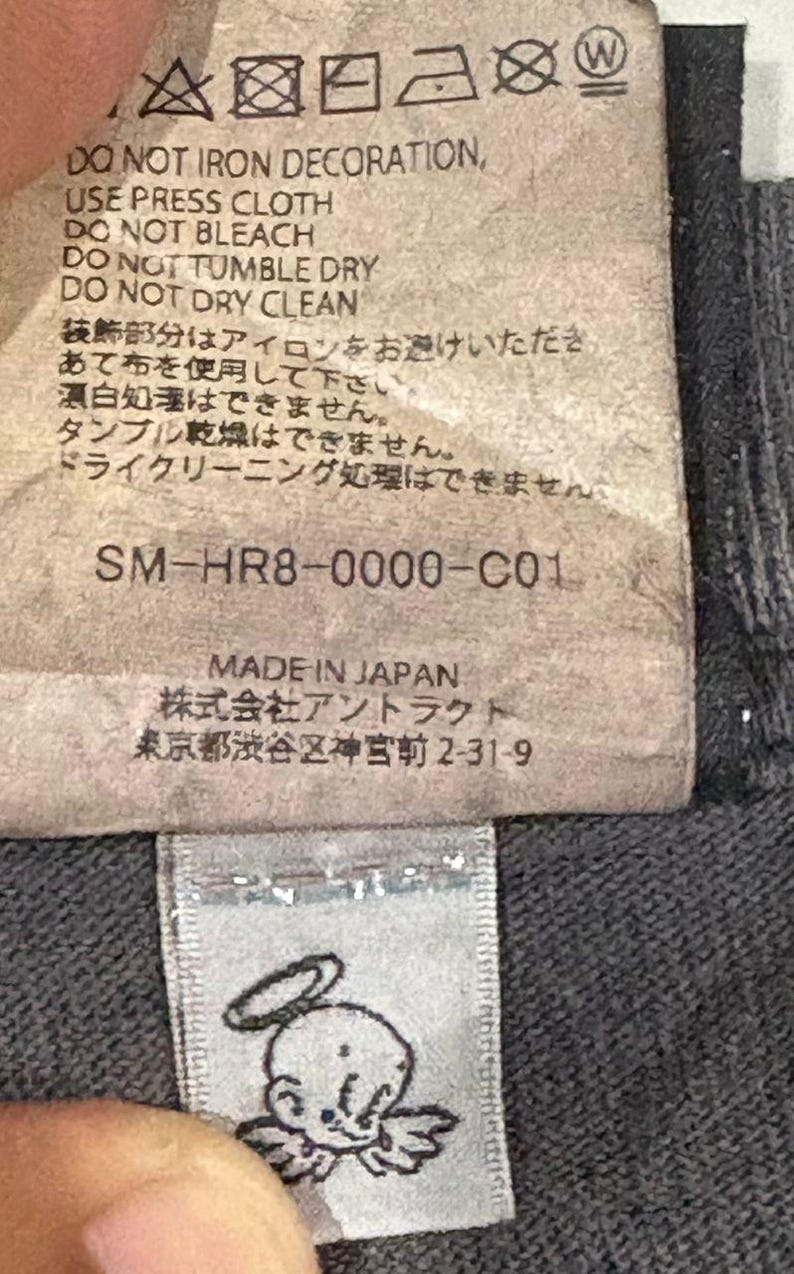 May include: A rectangular fabric care label with black text on a light background. It includes laundry symbols, instructions like DO NOT IRON DECORATION, the code SM-HR8-0000-C01, and MADE IN JAPAN. Below is a small white tag with a black line drawing of an angel.
