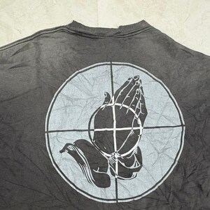 May include: A charcoal gray t-shirt featuring a large, light gray circular graphic on the back. The design depicts a pair of hands pressed together in a prayer position, overlaid with a crosshair pattern that divides the circle into four quadrants.