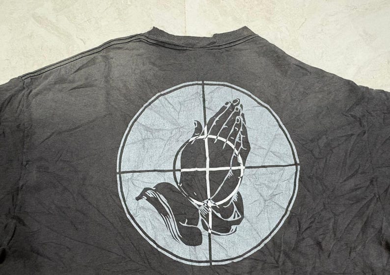 May include: A charcoal gray t-shirt featuring a large, light gray circular graphic on the back. The design depicts a pair of hands pressed together in a prayer position, overlaid with a crosshair pattern that divides the circle into four quadrants.