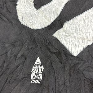 May include: A black t-shirt featuring a large, white, cracked-texture graphic at the top and a smaller white logo below. The smaller logo depicts a triangle with an eye, the text SAINT OF GOD, an infinity symbol, and the word ETERNAL underneath.
