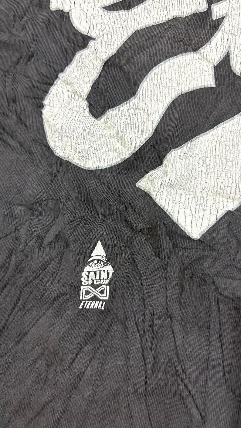 May include: A black t-shirt featuring a large, white, cracked-texture graphic at the top and a smaller white logo below. The smaller logo depicts a triangle with an eye, the text SAINT OF GOD, an infinity symbol, and the word ETERNAL underneath.