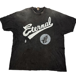 May include: A black short-sleeve t-shirt featuring the word Eternal in large white script lettering across the chest. Below the text is a circular graphic containing a pair of praying hands inside a crosshair target symbol and a small logo.