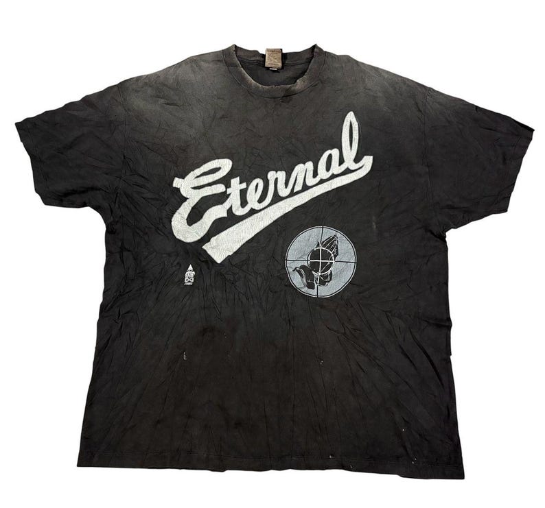 May include: A black short-sleeve t-shirt featuring the word Eternal in large white script lettering across the chest. Below the text is a circular graphic containing a pair of praying hands inside a crosshair target symbol and a small logo.