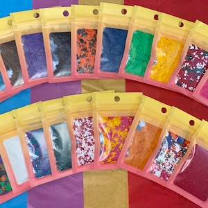 K&ouml;nnte beinhalten: Eine Sammlung kleiner, klarer Plastikt&uuml;ten, gef&uuml;llt mit bunten, unregelm&auml;&szlig;ig geformten Flocken. Die Flocken variieren in der Farbe, darunter Gelb, Blau, Orange und Lila. Jede T&uuml;te ist versiegelt und hat einen gelben Deckel.