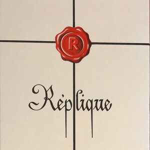 May include: A white box with a red wax seal with the letter "R" inside. The box has black lines and the text "Replique", "Eau de Toilette", "Raphael Paris", and "Net Cont 1.7 Fl. Ozs."