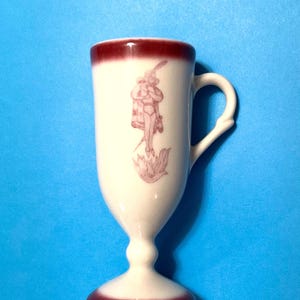May include: A white ceramic goblet-shaped mug with a handle, featuring a maroon rim and base. A maroon illustration of a figure with a hat and cape is on the front. The mug has a pedestal base and is set against a blue background.