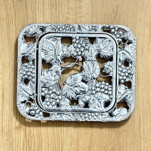 May include: A rectangular silver-colored metal trivet featuring an intricate embossed design of grape clusters and vine leaves. The frame has rounded corners and a repeating pattern of grapes and foliage that extends across the entire surface.