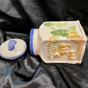 May include: White ceramic cookie jar with a blue trim and a whimsical design featuring a green forest scene with two deer. The lid has a blue handle.
