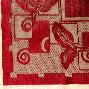 May include: Close-up of a red and beige fabric with a repeating pattern of leaves and geometric shapes. The fabric has a textured, woven look.