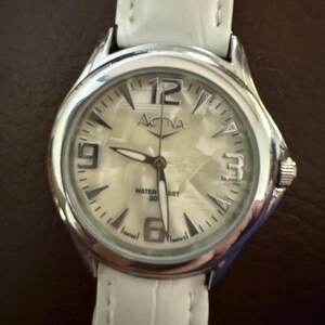 Op de afbeelding: Een zilveren Activa horloge met een witte leren band. De wijzerplaat van het horloge is lichtcr&egrave;me van kleur met zwarte cijfers en wijzers. Het horloge is waterdicht tot 30 meter.