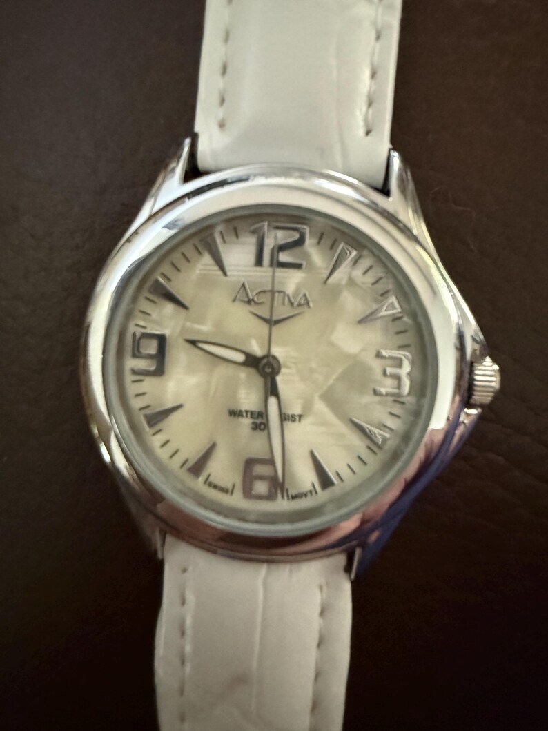 Op de afbeelding: Een zilveren Activa horloge met een witte leren band. De wijzerplaat van het horloge is lichtcr&egrave;me van kleur met zwarte cijfers en wijzers. Het horloge is waterdicht tot 30 meter.
