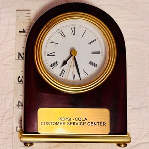 May include: A dark wood clock with a gold trim and a white face. The clock has Roman numerals and a gold second hand. The clock is sitting on a gold base with a plaque that reads "Pepsi-Cola Customer Service Center".