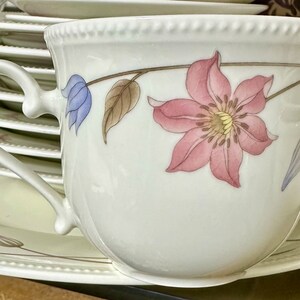 May include: A white porcelain teacup with a floral design featuring a pink flower with a yellow center, a blue flower, and brown leaves. The cup has a handle and is sitting on a white saucer.
