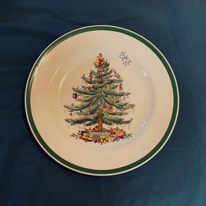 May include: A white ceramic plate with a green border. The plate features a detailed illustration of a decorated Christmas tree with ornaments, gifts, and Santa Claus at the top. A festive holiday design.