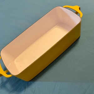 May include: A rectangular yellow baking dish with a white interior. The dish has two yellow handles and is set against a blue background. This cookware is ideal for baking casseroles, bread, and other dishes.