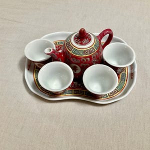 May include: A ceramic tea set featuring a red teapot with a lid and three matching white-interior cups arranged on a white, wavy-edged tray. The teapot and tray are decorated with intricate red, green, and gold geometric and floral patterns.