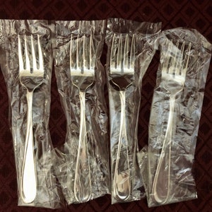 May include: Four silver forks are displayed in individual plastic packaging. The forks are all the same design and have a polished finish.