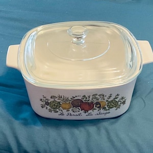 May include: A square, off-white casserole dish with a clear glass lid and handles. The dish features a colorful botanical design with fruits and vegetables, and text that reads "Le Persil, La Sauge".