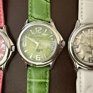 Op de afbeelding: Drie Activa-horloges met zilveren kasten en leren bandjes. De horloges hebben witte wijzerplaten met zwarte cijfers en wijzers. De bandjes zijn roze, groen en wit. De wijzerplaten tonen de merknaam en het nummer 12.