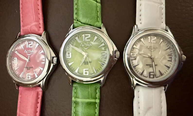 Op de afbeelding: Drie Activa-horloges met zilveren kasten en leren bandjes. De horloges hebben witte wijzerplaten met zwarte cijfers en wijzers. De bandjes zijn roze, groen en wit. De wijzerplaten tonen de merknaam en het nummer 12.
