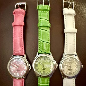 Op de afbeelding: Drie analoge horloges met zilverkleurige kasten en leren bandjes in roze, groen en wit. De wijzerplaten tonen de merknaam "Activa" en de cijfers 3, 6, 9 en 12. De bandjes hebben een textuur met krokodillenpatroon.