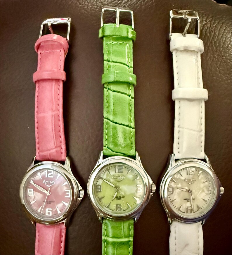 Op de afbeelding: Drie analoge horloges met zilverkleurige kasten en leren bandjes in roze, groen en wit. De wijzerplaten tonen de merknaam "Activa" en de cijfers 3, 6, 9 en 12. De bandjes hebben een textuur met krokodillenpatroon.