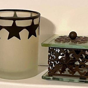 May include: Two decorative candle holders, one with a frosted glass cylinder and a metal rim with black star cutouts, the other with a metal frame with a star design and a glass base.