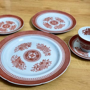 May include: A set of vintage porcelain dishware with a floral design. The set includes four plates of varying sizes, a teacup, and a saucer. The plates and cup have a white background with a red floral pattern and a red border.