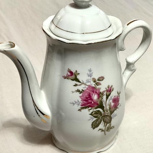 May include: A white porcelain teapot featuring a delicate floral pattern of pink roses and green leaves. The teapot has a scalloped rim, a curved handle, a spout, and a matching lid with a gold-painted finial and gold trim along the edges.