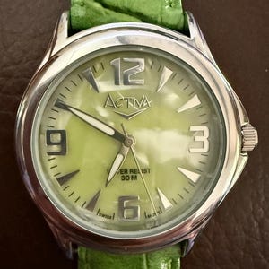 Op de afbeelding: Zilverkleurig ACTIVA horloge met een lichtgroene wijzerplaat en een groene leren band. De wijzerplaat heeft grote zilveren cijfers op 12, 3, 6 en 9 uur. De merknaam "ACTIVA" is prominent aanwezig.
