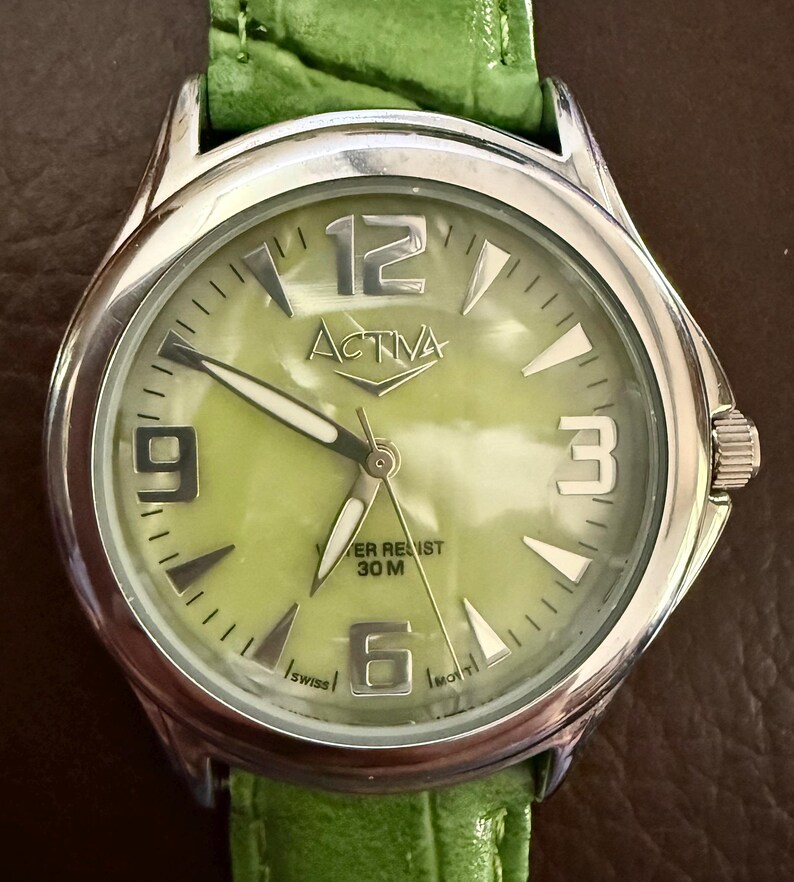 Op de afbeelding: Zilverkleurig ACTIVA horloge met een lichtgroene wijzerplaat en een groene leren band. De wijzerplaat heeft grote zilveren cijfers op 12, 3, 6 en 9 uur. De merknaam "ACTIVA" is prominent aanwezig.