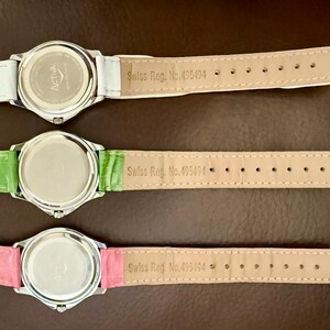 Op de afbeelding: Drie horloges met zilverkleurige kasten en leren bandjes in wit, groen en roze. De achterkanten van de horloges zijn zichtbaar, met de merknaam "ACTIVA" en de tekst "Swiss Reg. No. 495494" op de bandjes.